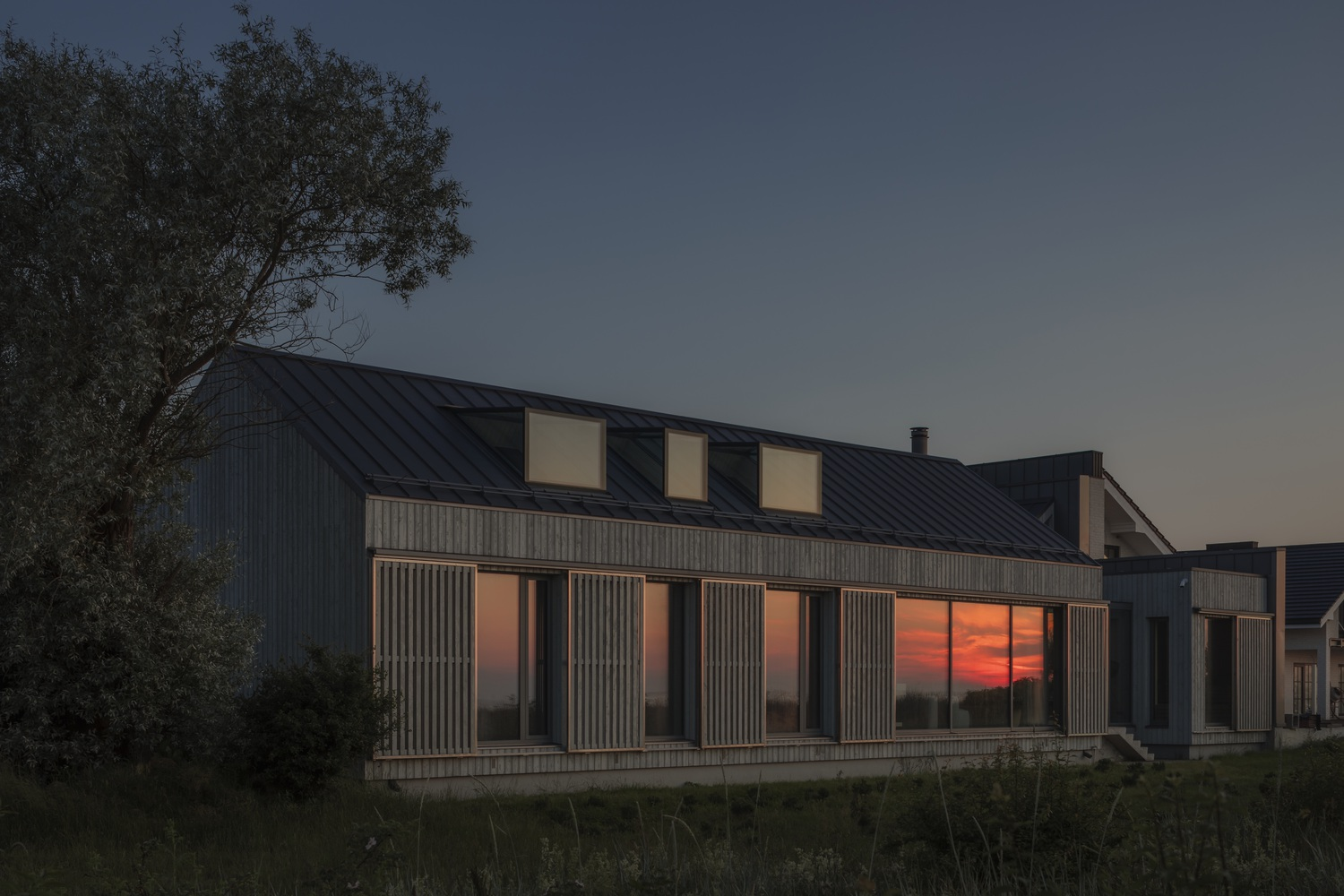 Vertical timber screens fronting glass walls reflecting sunset with tree in foreground at twilight