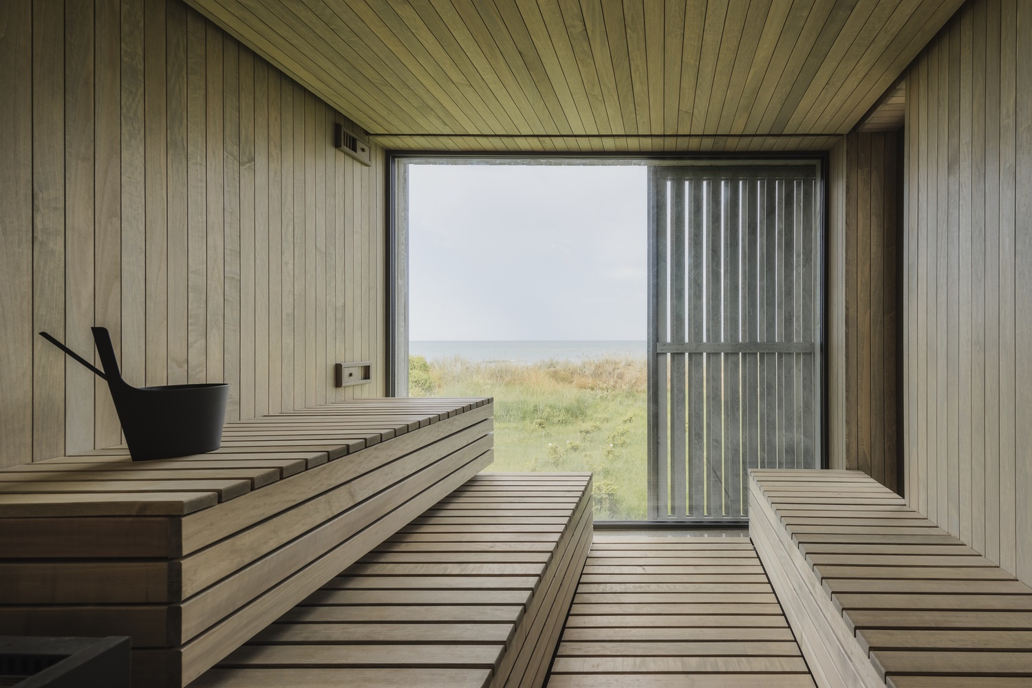 Tiered timber sauna benches surrounding a glazed opening with coastal grassland and misty sky beyond