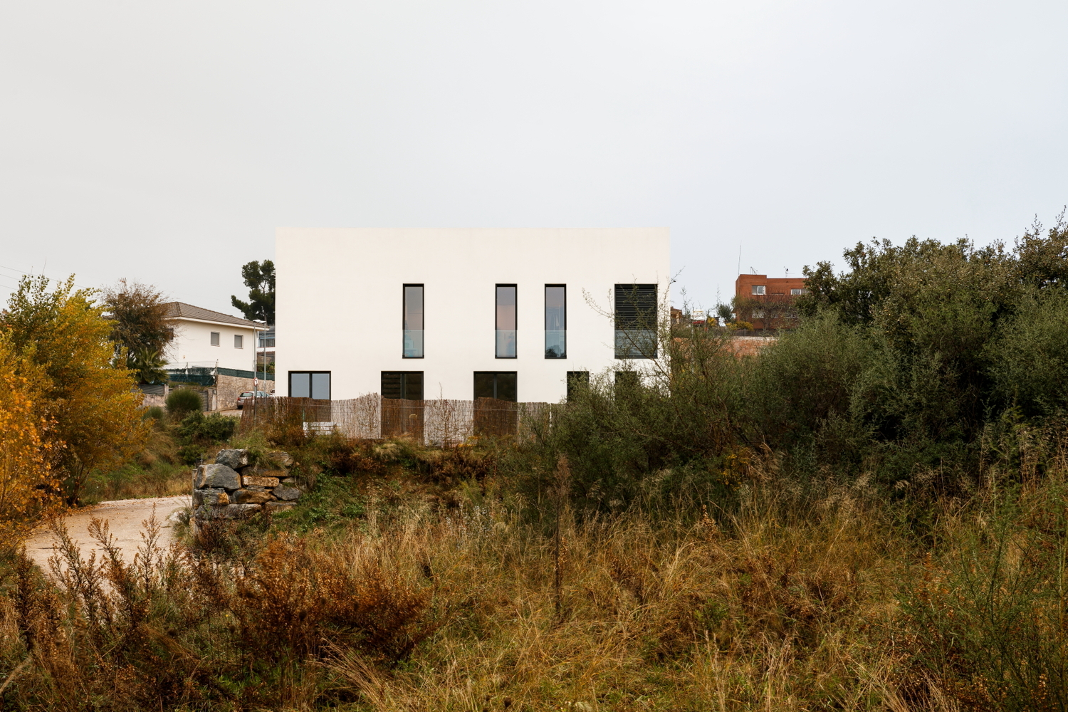 White rendered facade with vertical window openings viewed across overgrown autumn grasses and shrubs