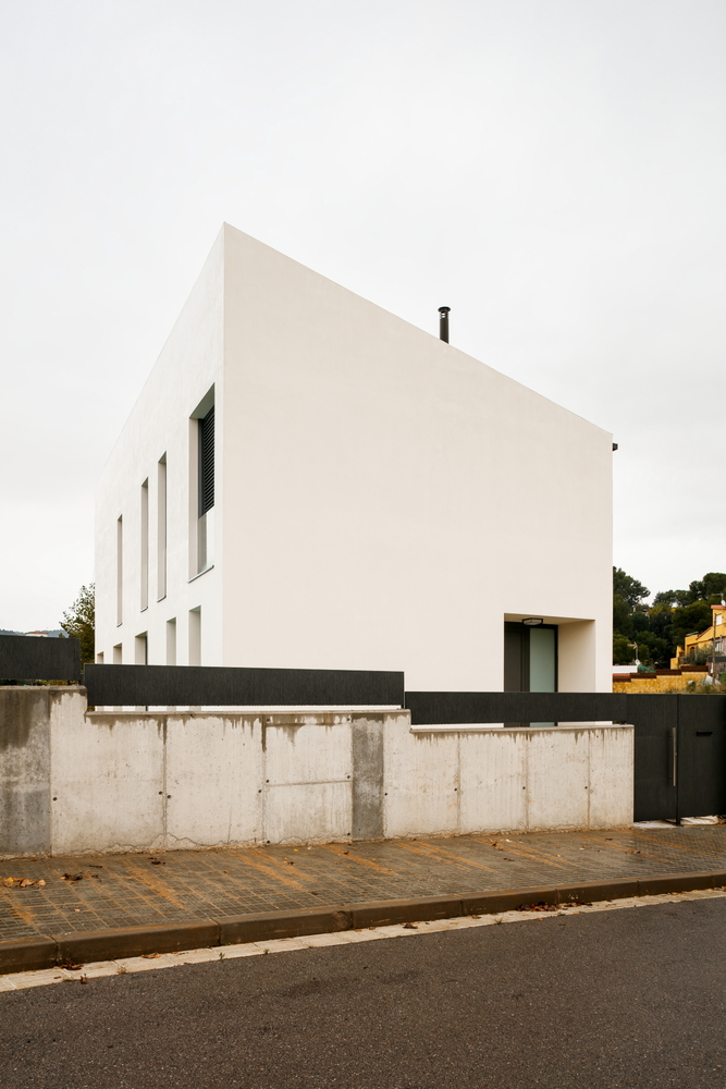 Street view of white stucco volume rising above exposed concrete retaining wall with dark metal gate