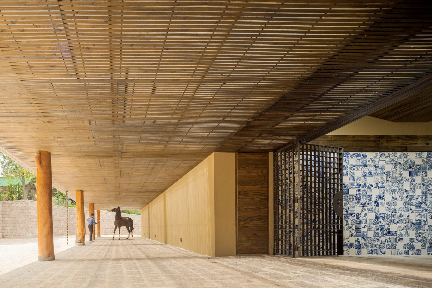 Covered colonnade with timber slat ceiling and blue tile mosaic wall beside a horse