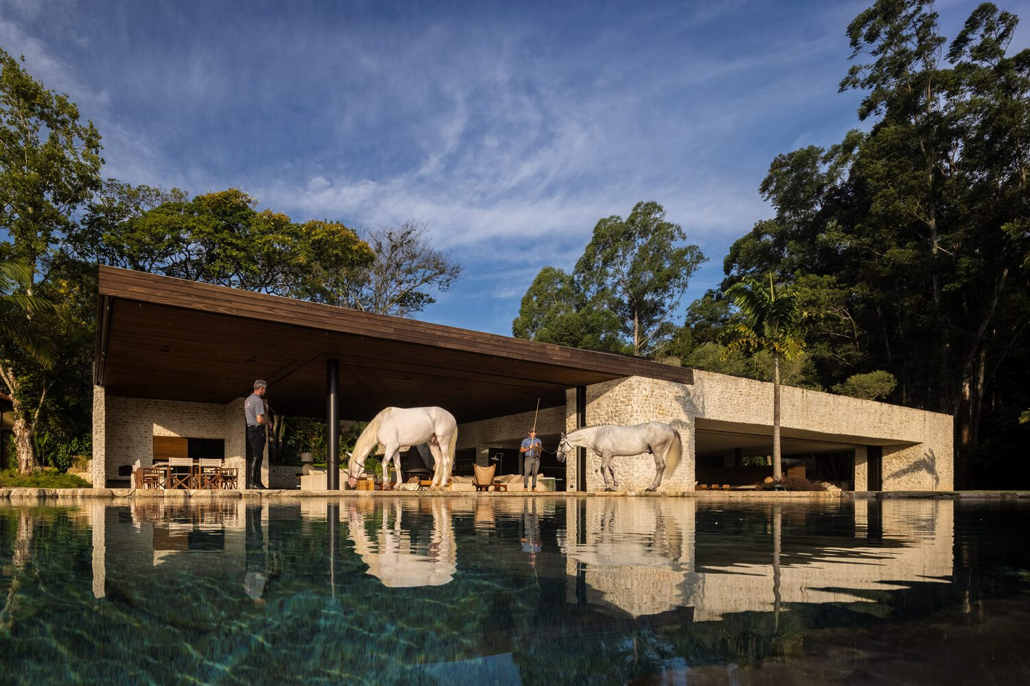Stone and timber pavilion reflected in pool with horses and figures under afternoon light