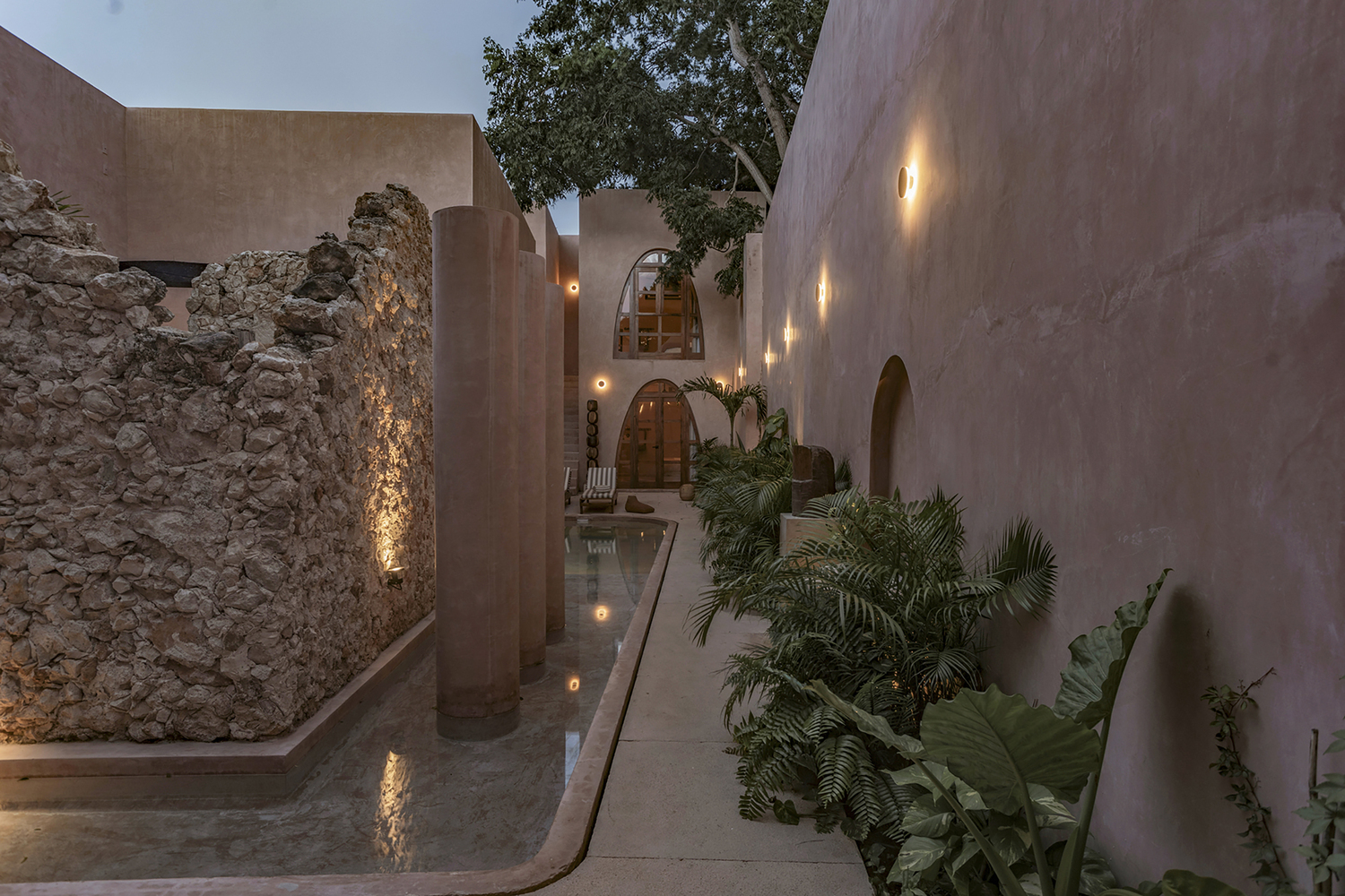 Courtyard passage with preserved stone ruins, rendered walls, arched openings and tropical plantings at dusk