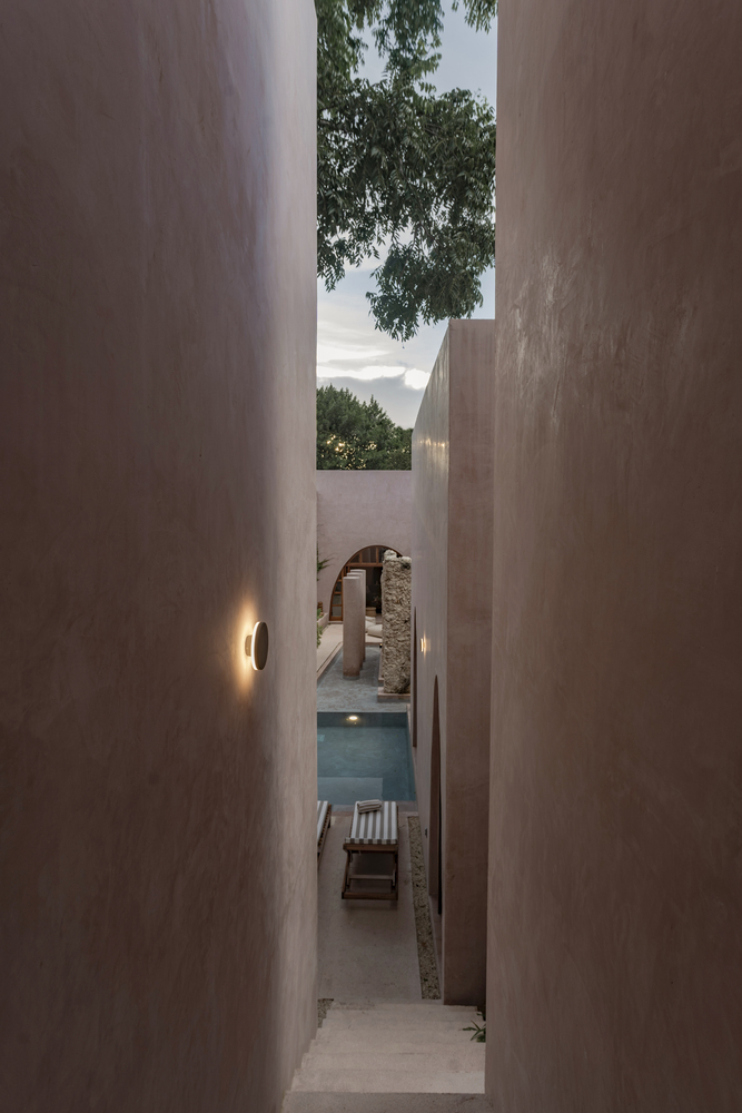 Narrow passage between plastered walls leading to a pool and preserved stone archway at twilight