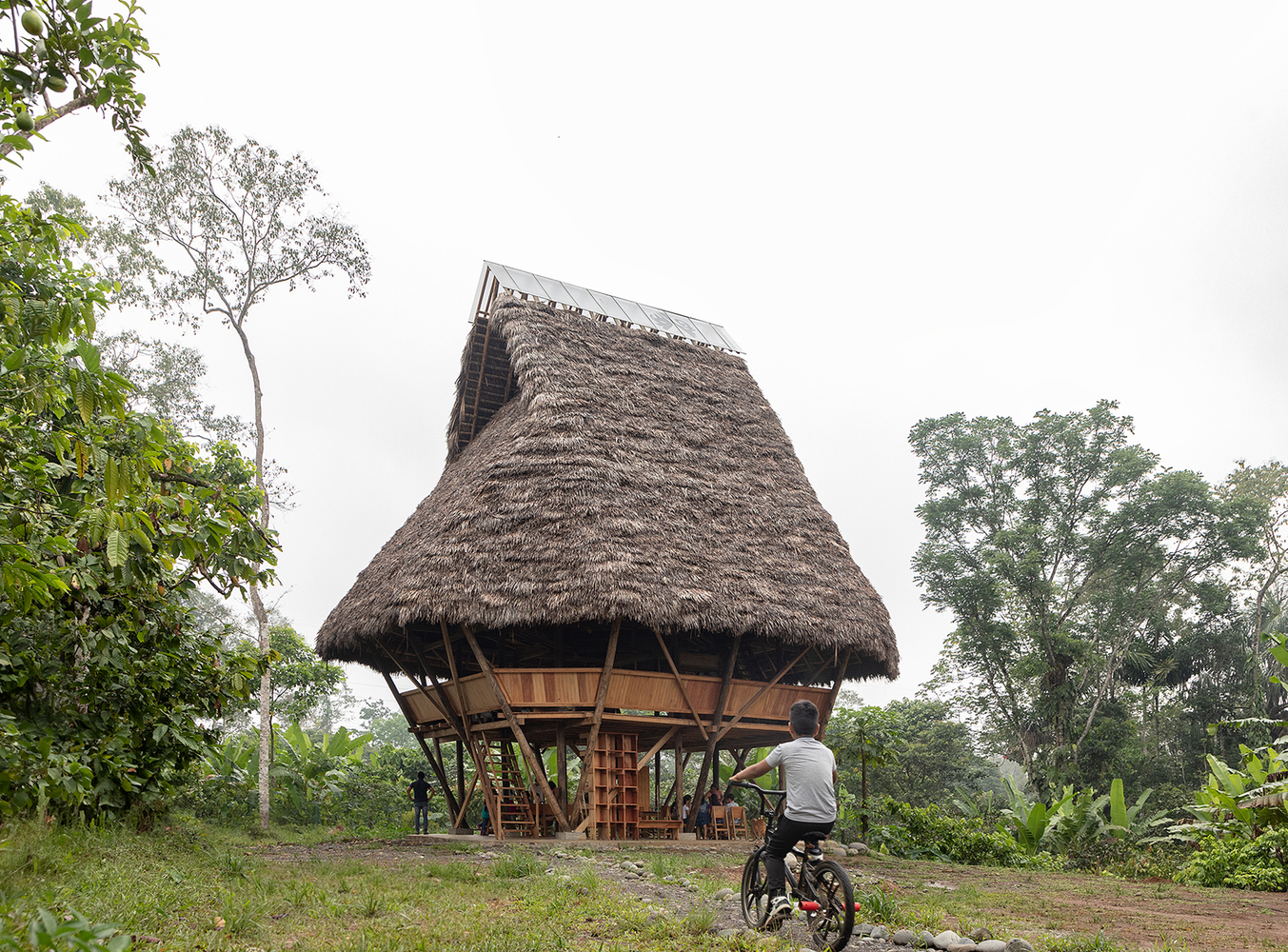 Elevated thatched roof pavilion on slender timber columns with a cyclist pausing on the dirt clearing below