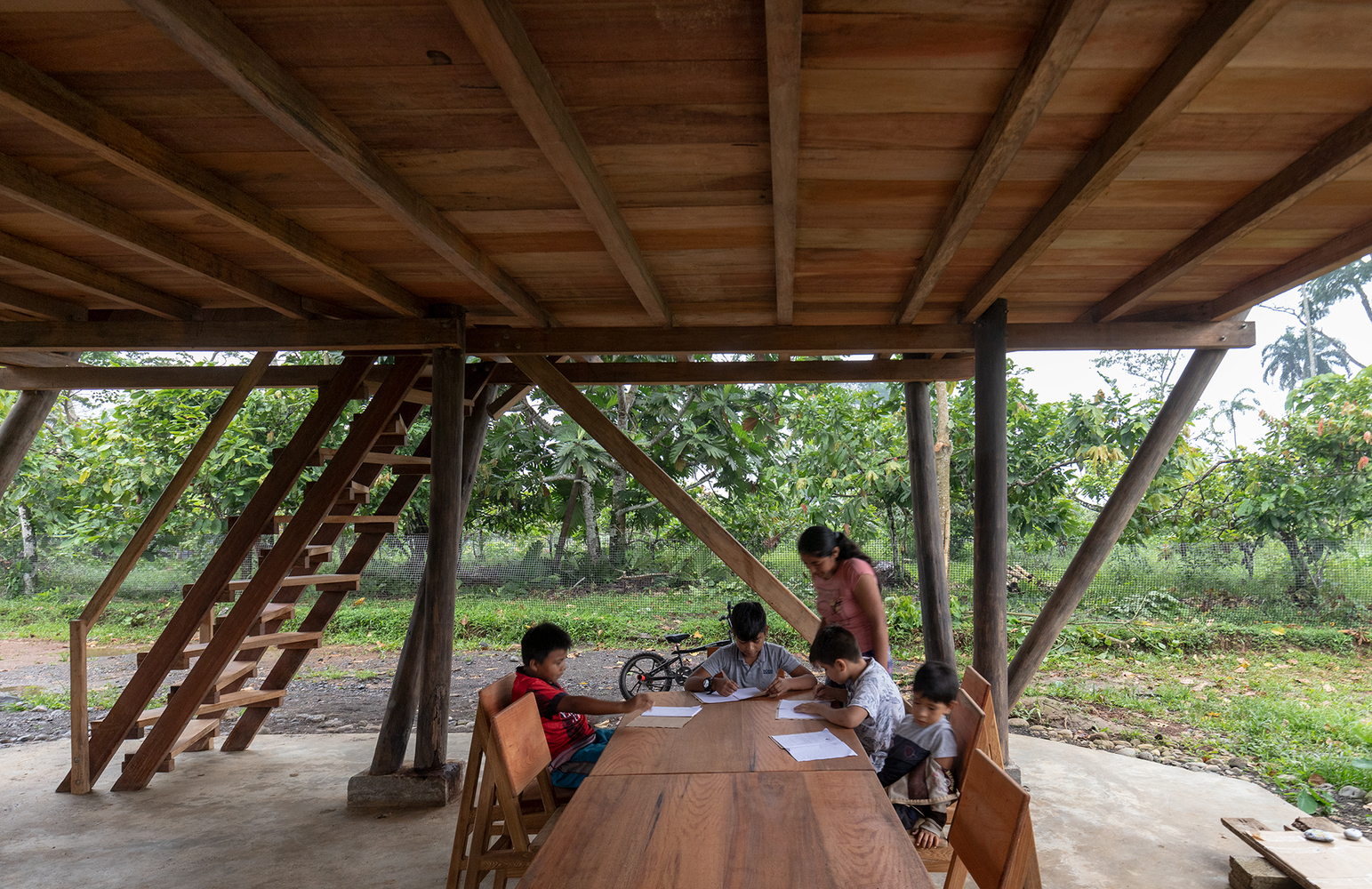 Ground-level pavilion with exposed timber beams and open sides where children gather at wooden benches