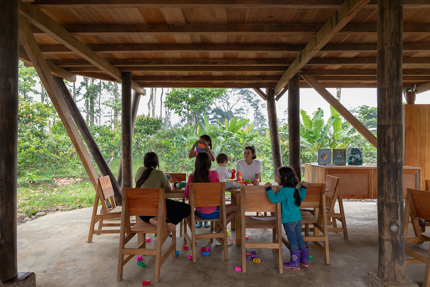 Children seated at timber tables under a covered platform framed by diagonal wooden struts
