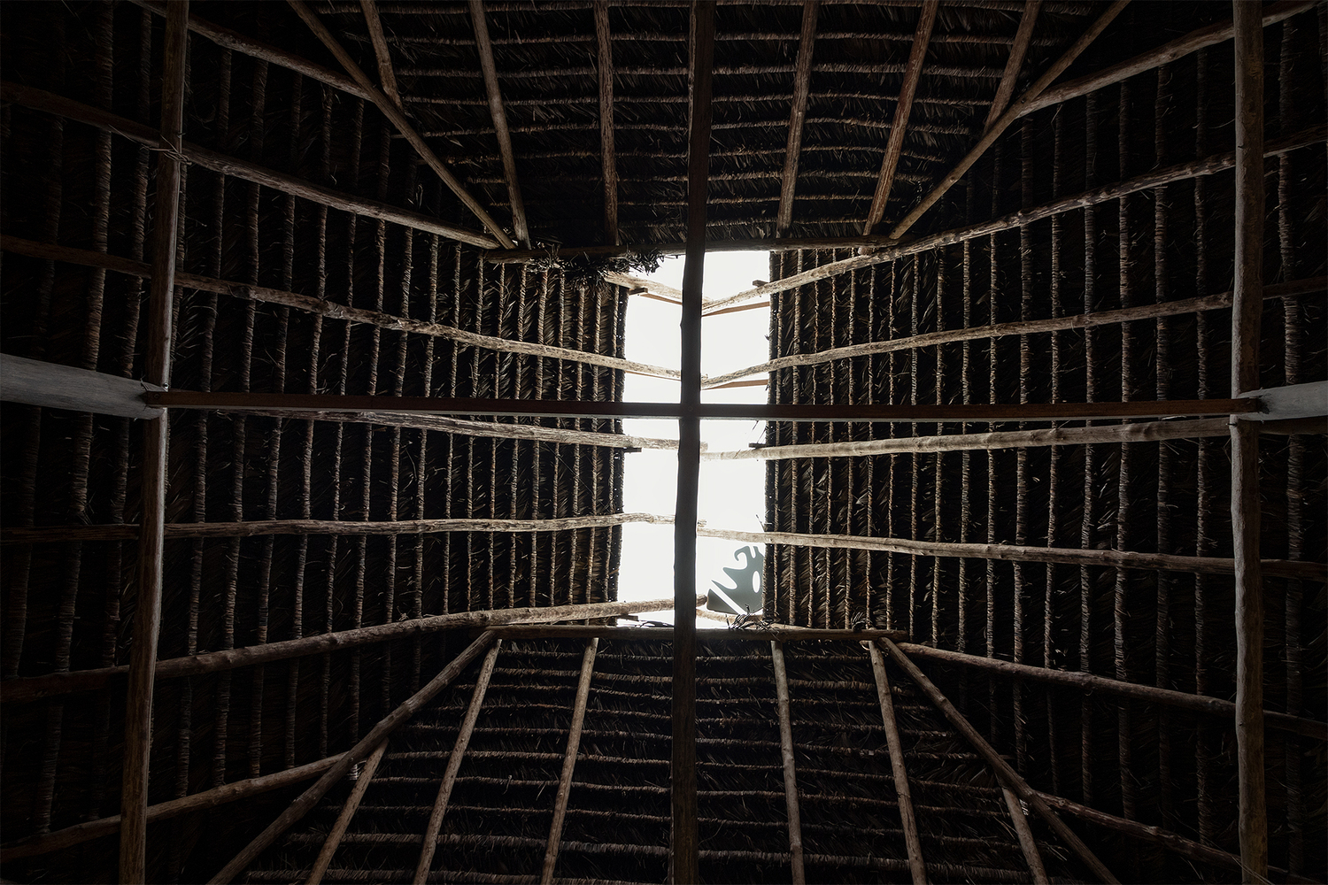 Upward view through the central timber roof structure with thatch infill and a silhouetted figure above