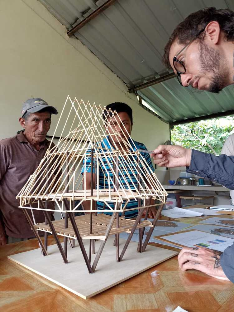 Three people examining a timber frame structural model under a corrugated metal roof in a workshop