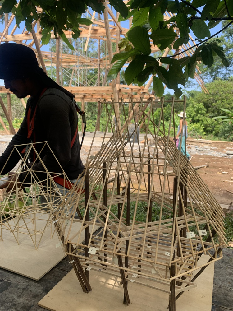 Bamboo structural model in foreground with full-scale timber frame construction visible beyond under tree canopy