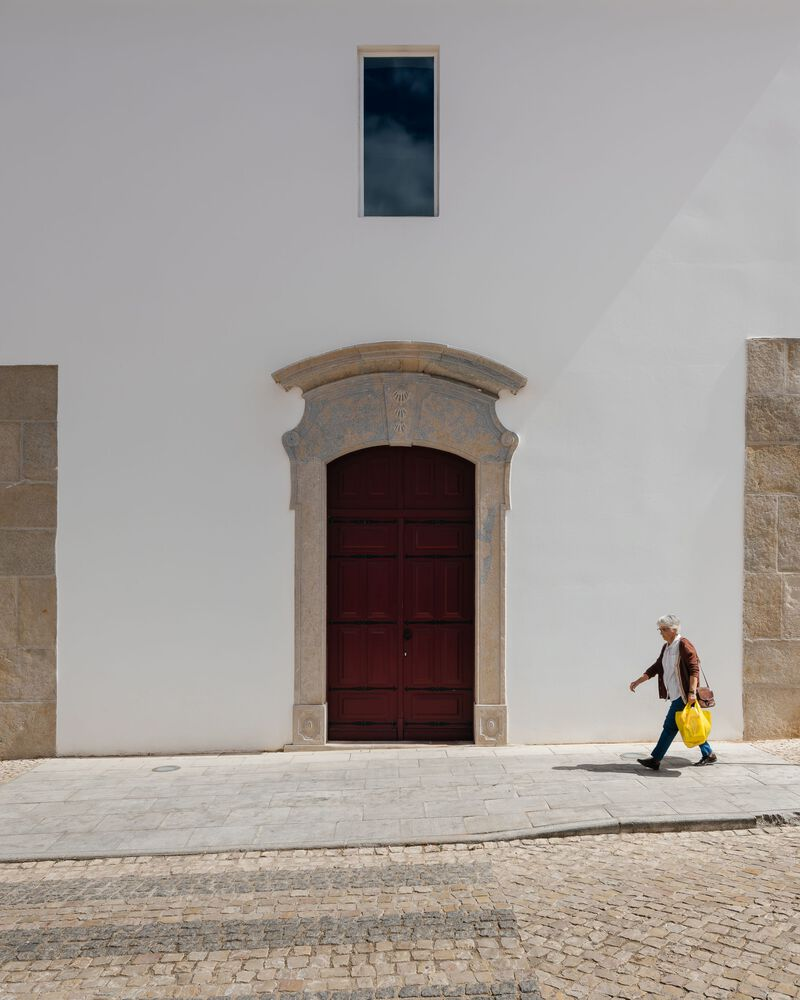 White stucco facade with stone-framed wooden door and narrow window as a pedestrian passes
