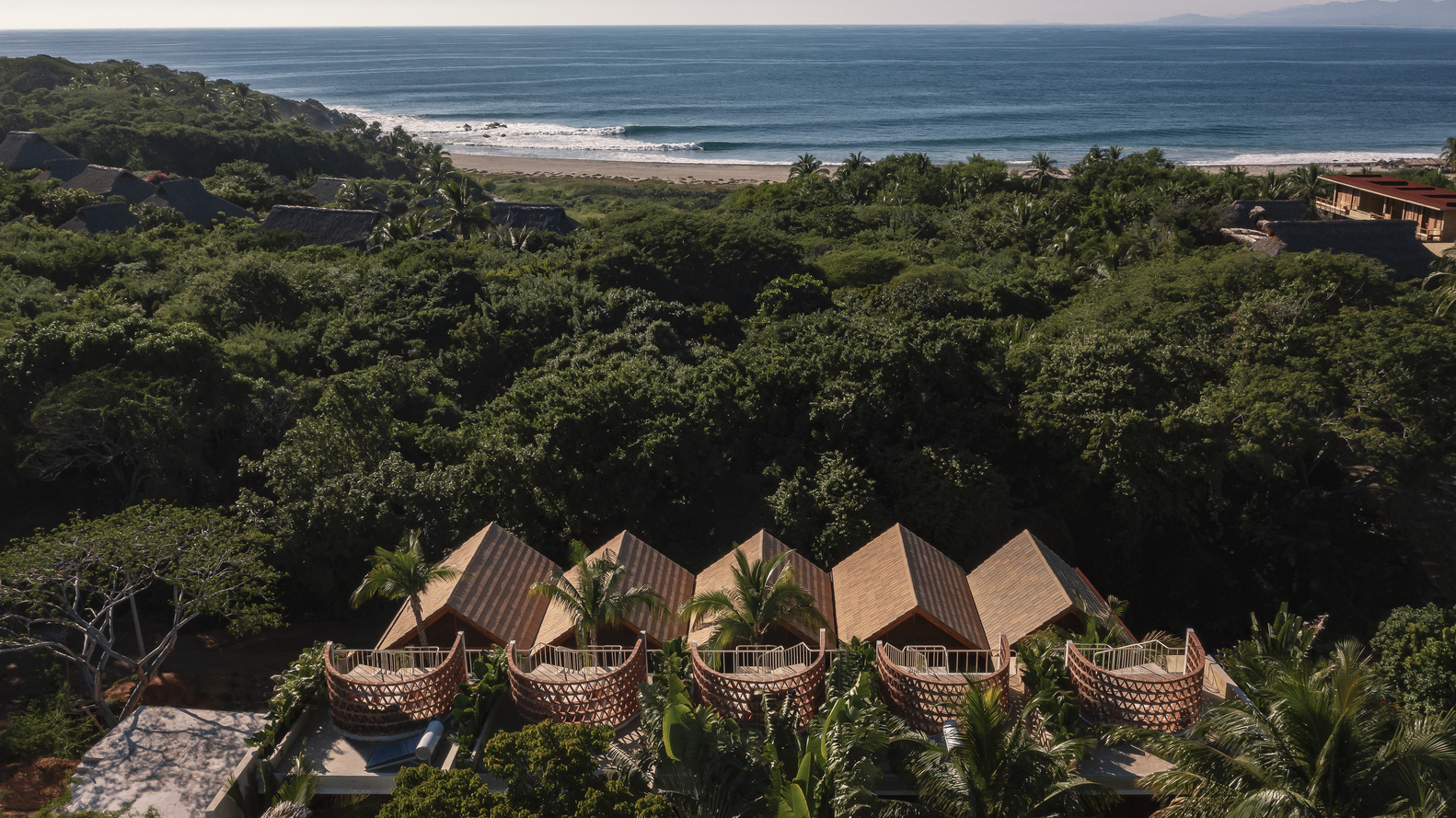 Distant view of the clustered pavilions with pitched roofs and perforated facades facing the ocean through forest canopy