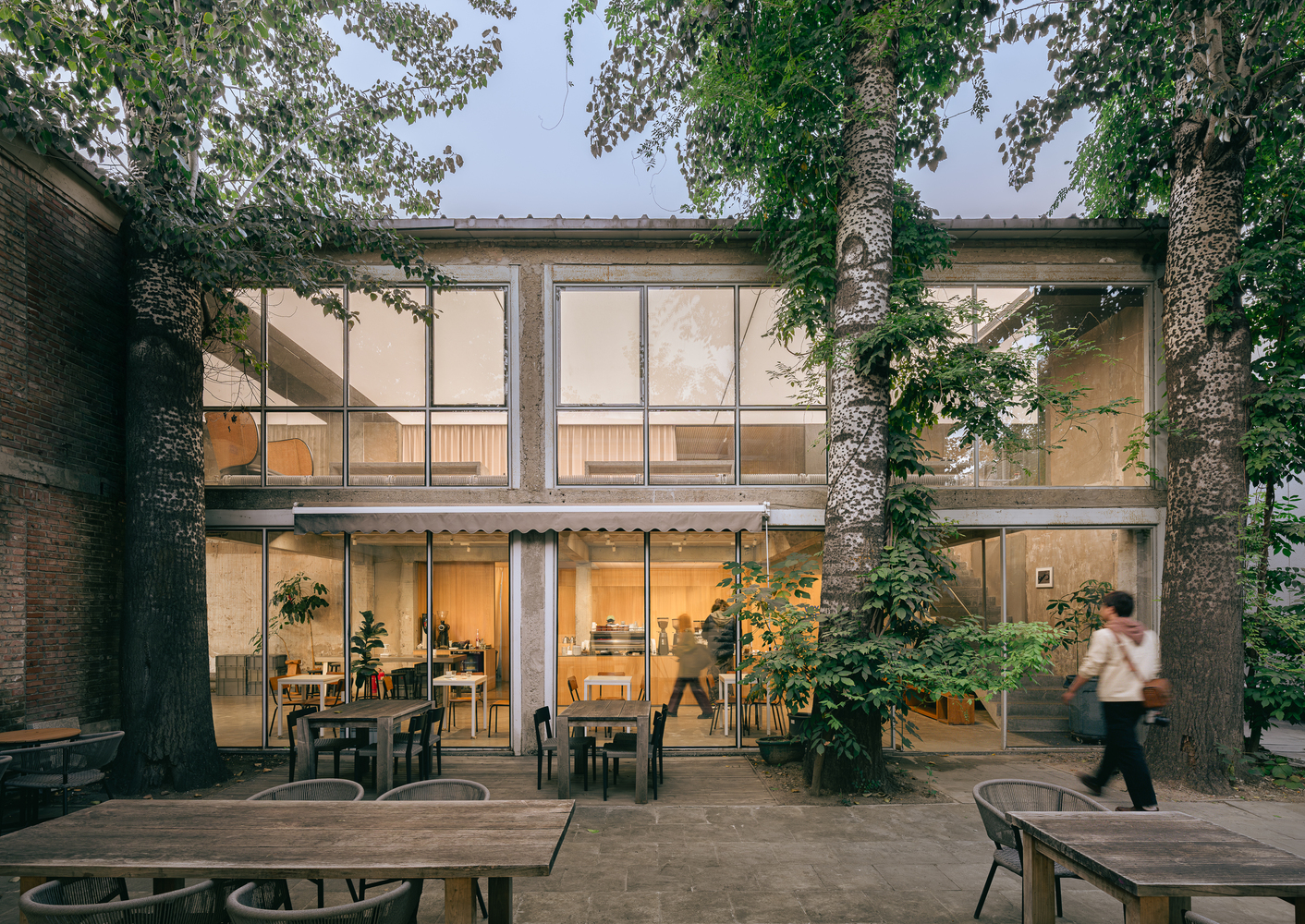 Two-story glass facade framed by concrete columns and mature trees in a courtyard at dusk