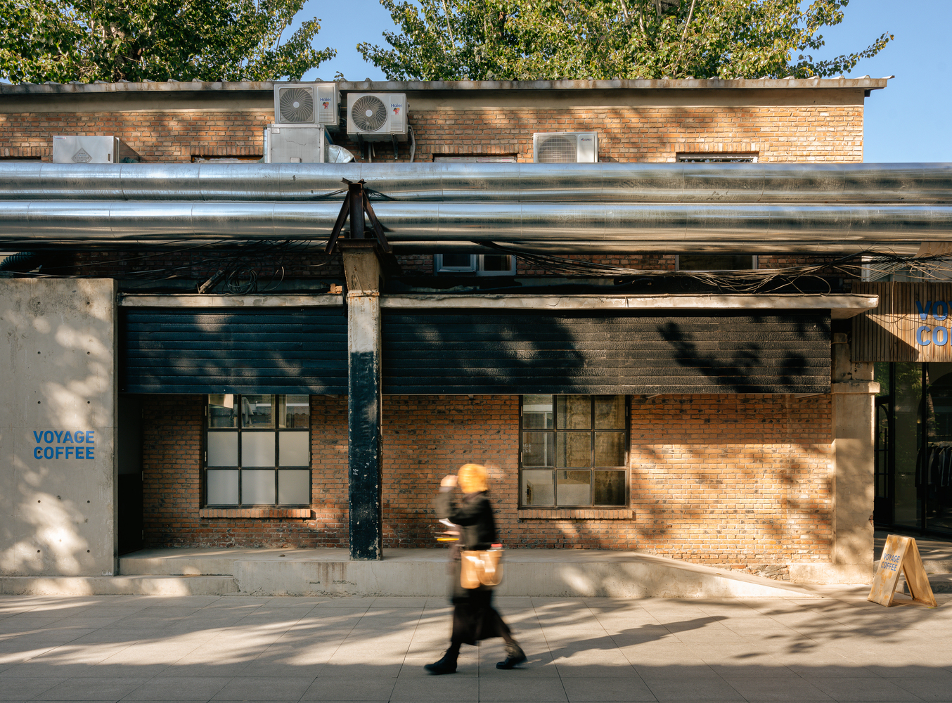 Brick facade with exposed mechanical units and a pedestrian passing beneath dappled tree shadows