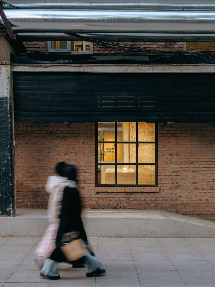 Exposed brick facade with steel-framed window and overhead ductwork as a figure passes by