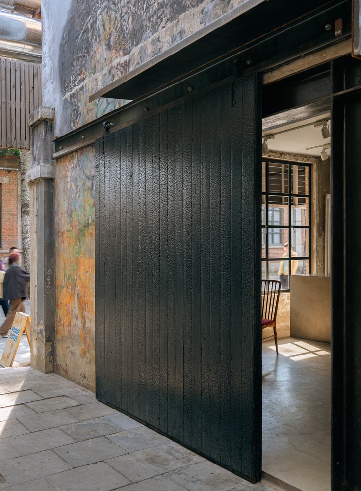 Charred timber sliding door partially open against distressed concrete wall with passerby in motion
