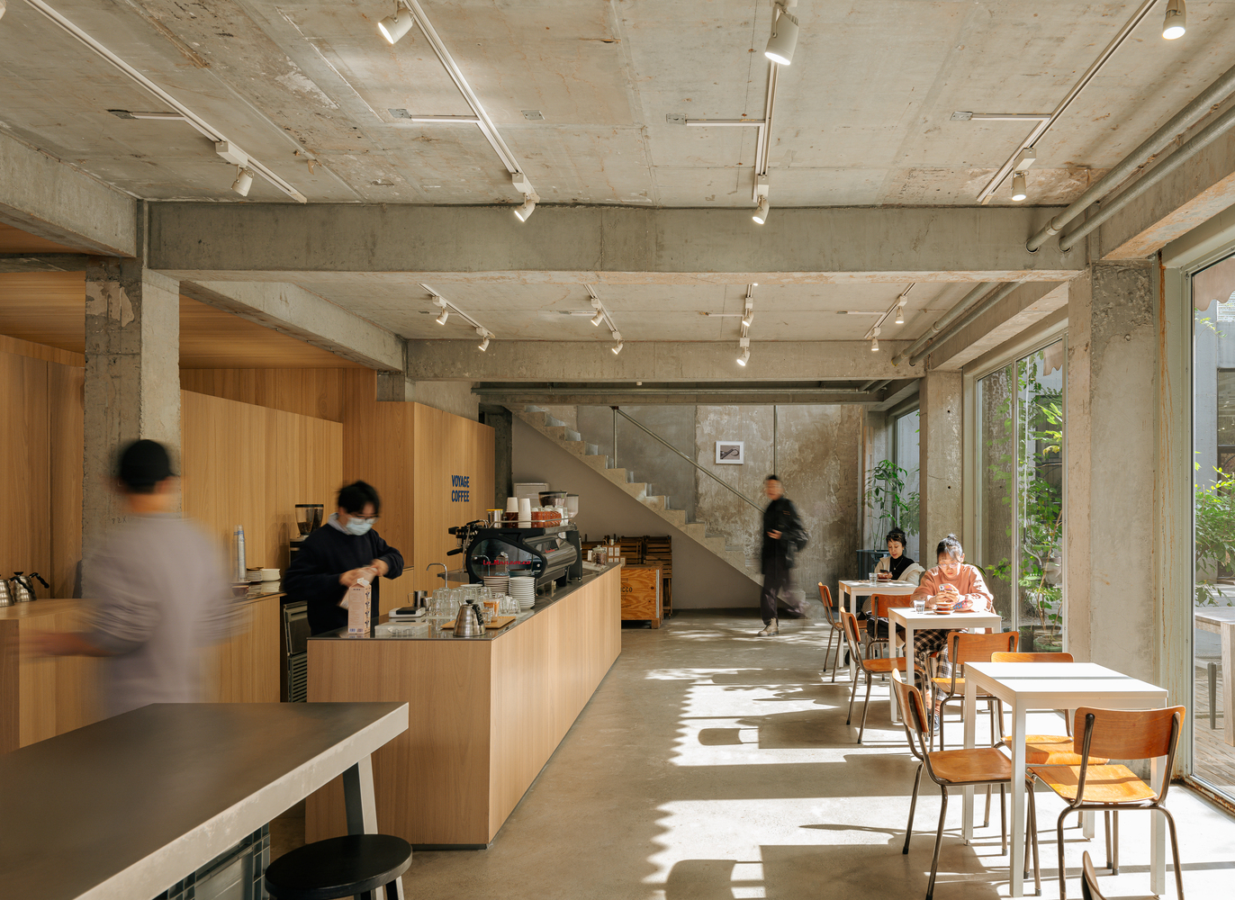 Cafe interior with exposed concrete ceiling and timber counter as patrons gather in dappled sunlight