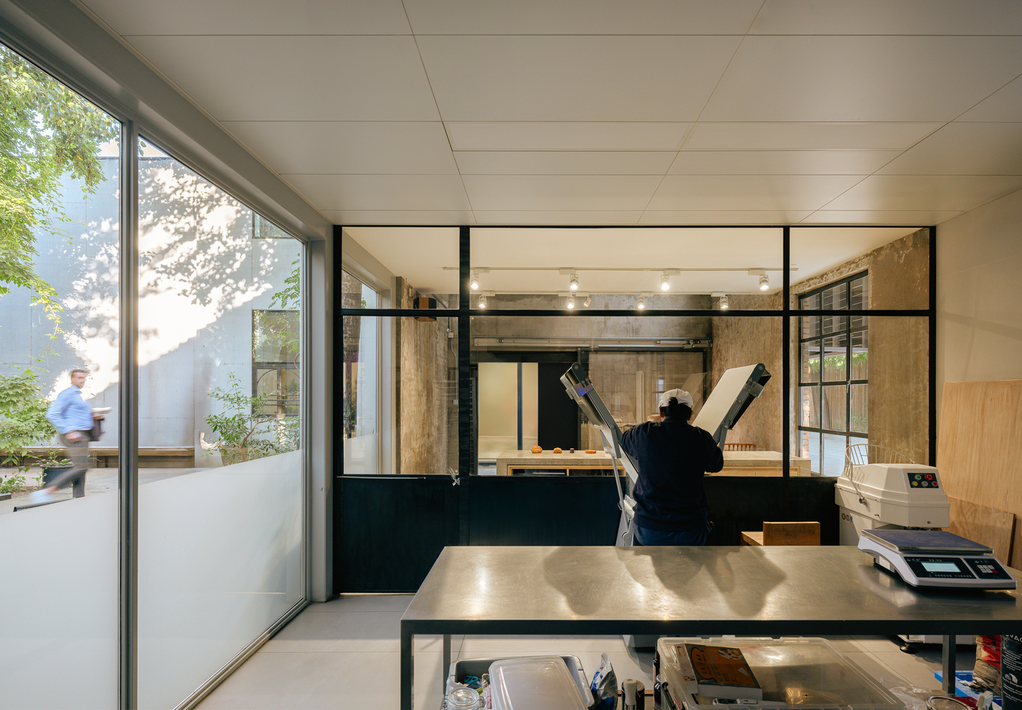 Workshop interior with steel worktable and floor-to-ceiling glazing opening to a planted courtyard