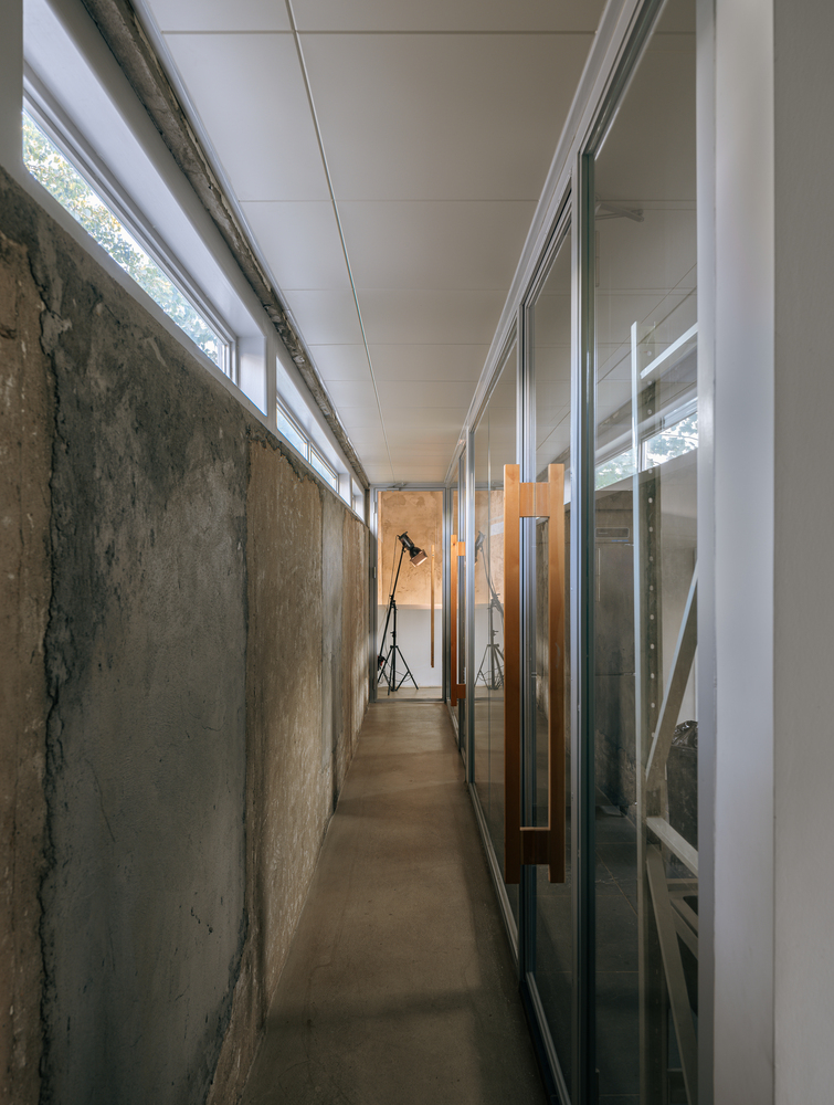 Narrow corridor with exposed concrete wall and glazed partitions beneath a ribbon of clerestory windows