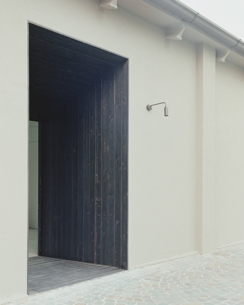 Exterior entrance with charred timber vertical cladding and pivoting door beside a pale rendered wall