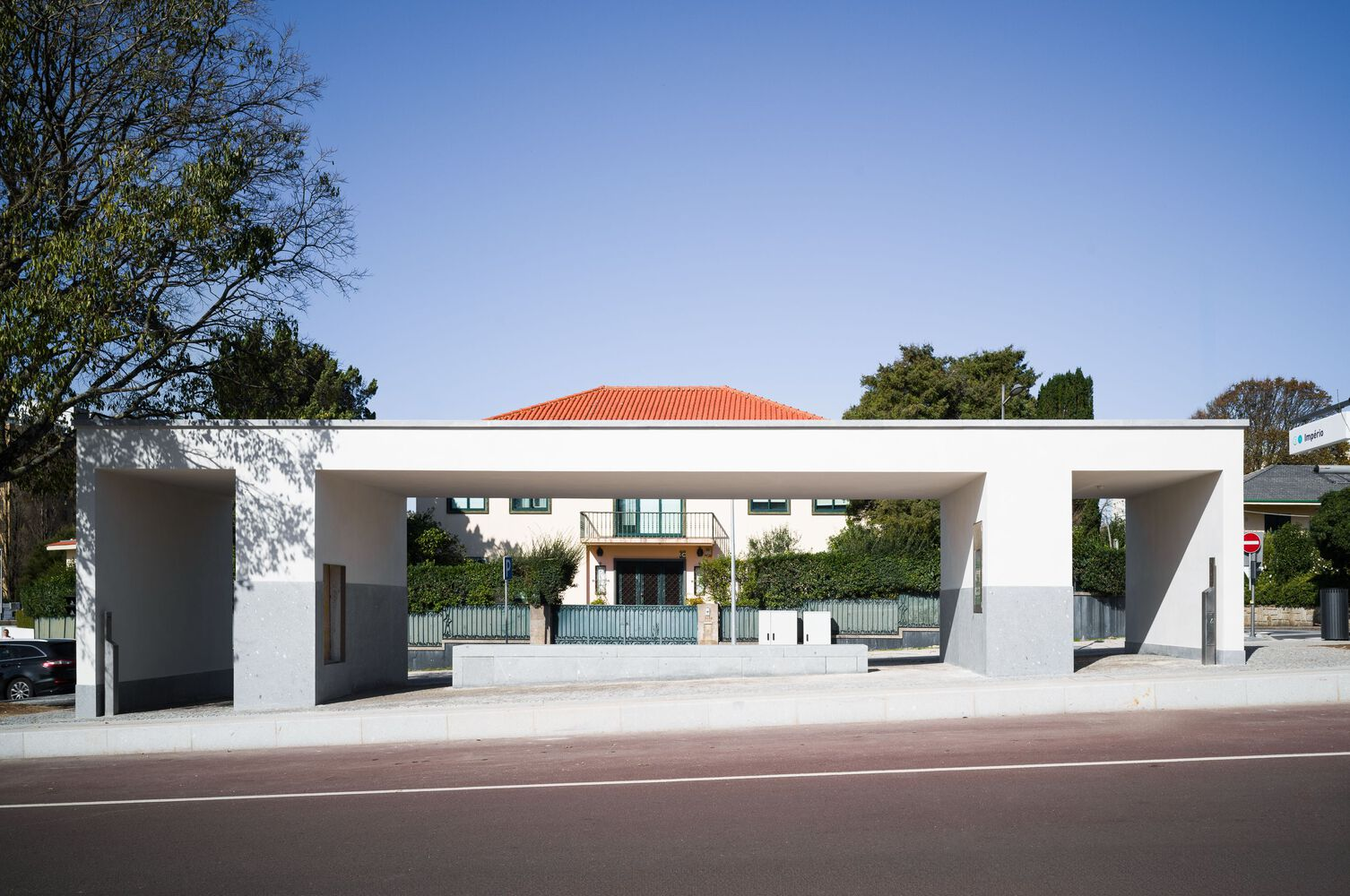 White concrete canopy structure with orange-tiled roof visible behind a tree-lined streetscape