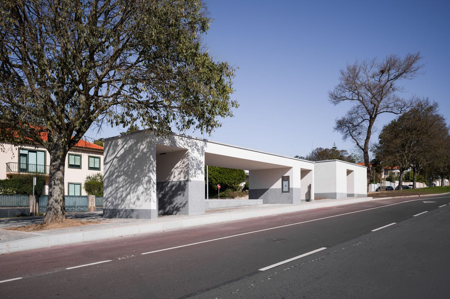 White concrete canopy and shelter structure casting tree shadows along a red paved roadway