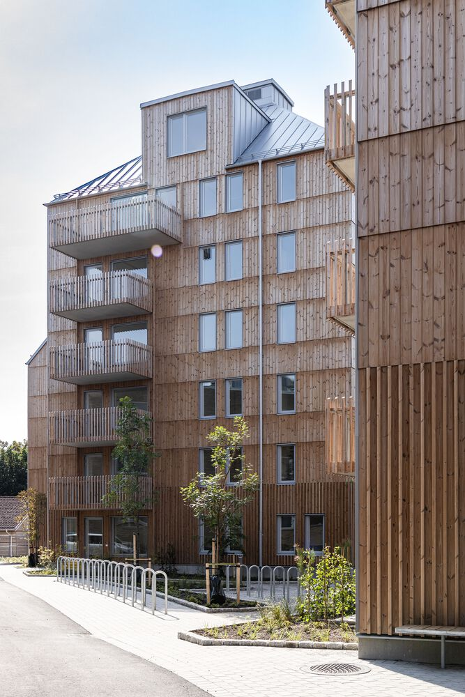 Timber-clad residential block with staggered balconies and metal roofs beside newly planted street trees
