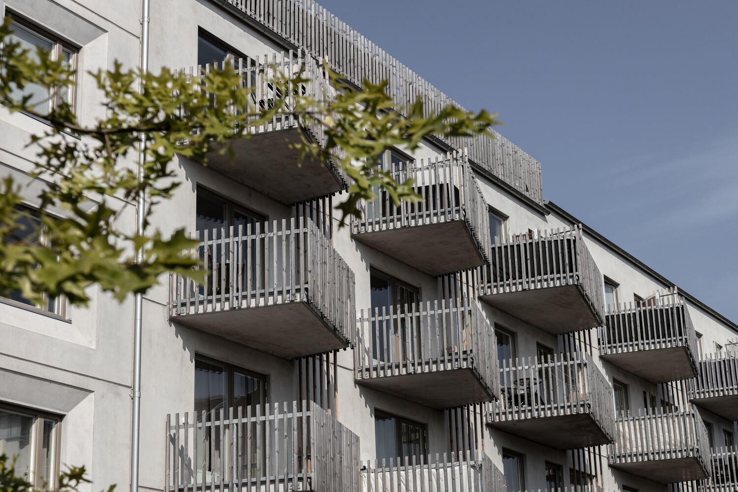 Facade with stacked concrete balconies and vertical white screening elements viewed through green foliage