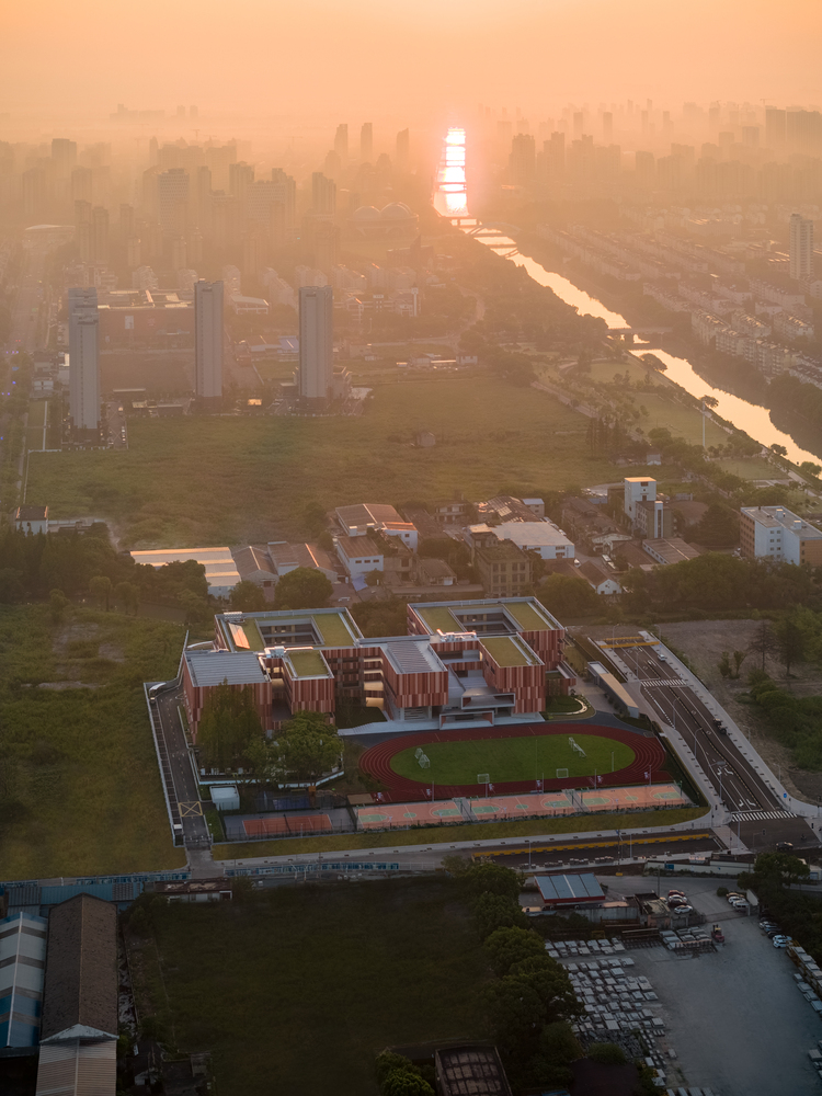 Aerial view of the school campus with vertical striped cladding and green rooftop sports field at golden hour