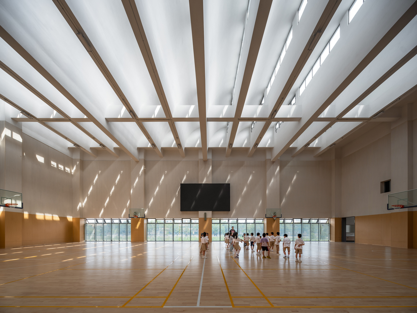 Interior sports hall with timber louvered ceiling casting diagonal shadows across students practicing on wooden floor