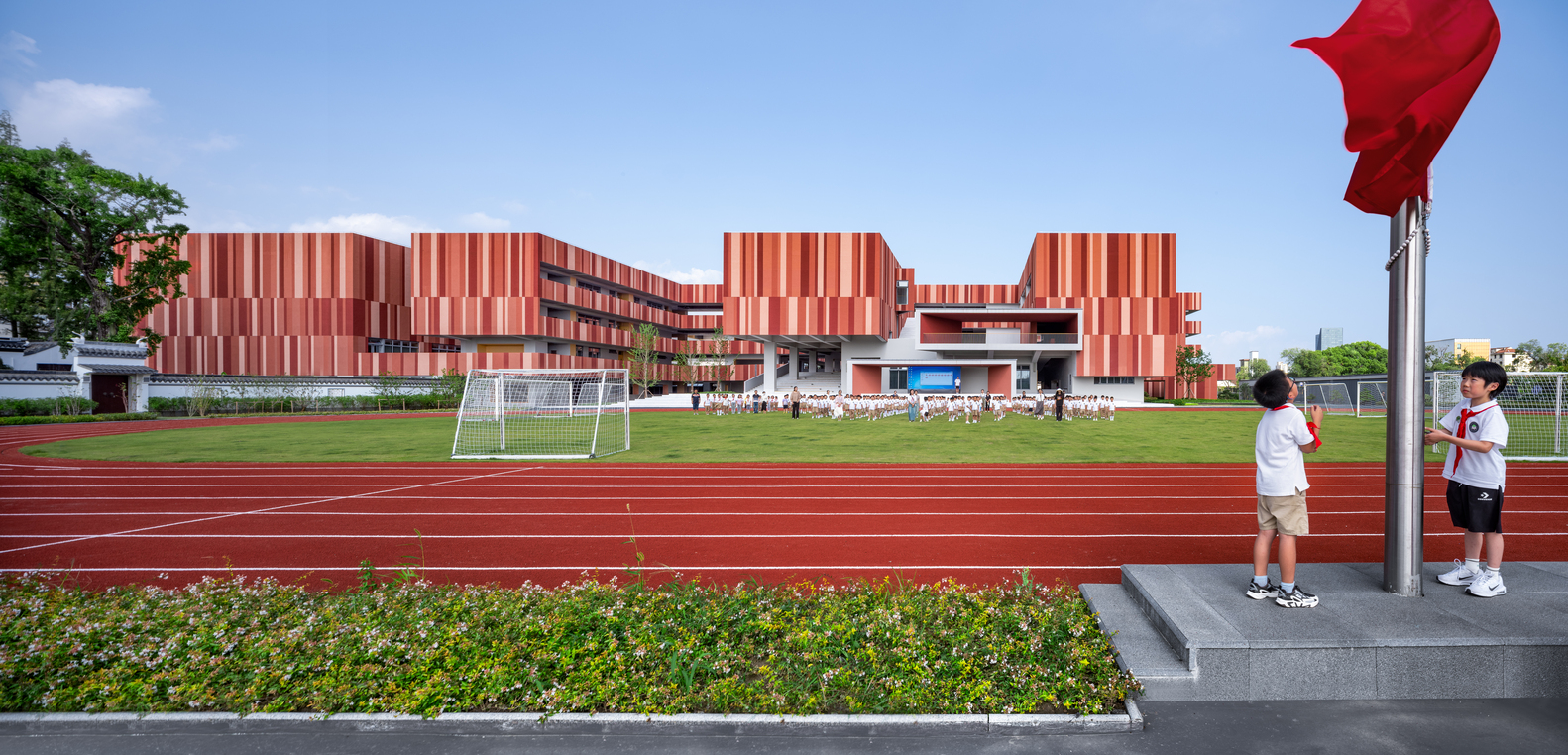View across the running track toward the striped facade with students and red flag in foreground