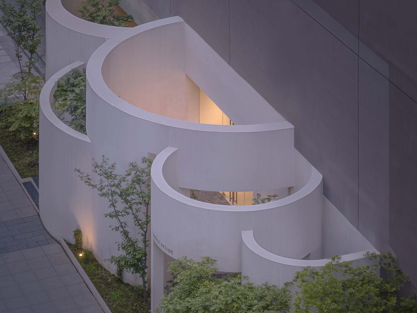 Aerial view of spiraling white volumes with planted terraces illuminated at dusk