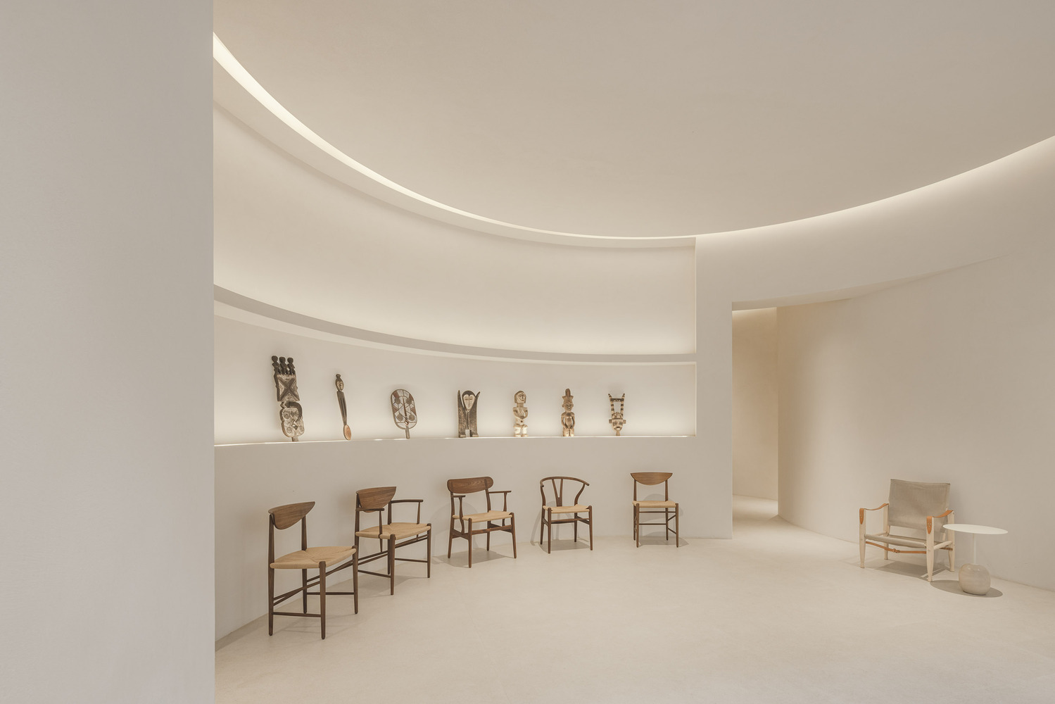 Curved display alcove with built-in shelving presenting wooden chairs and sculptural objects