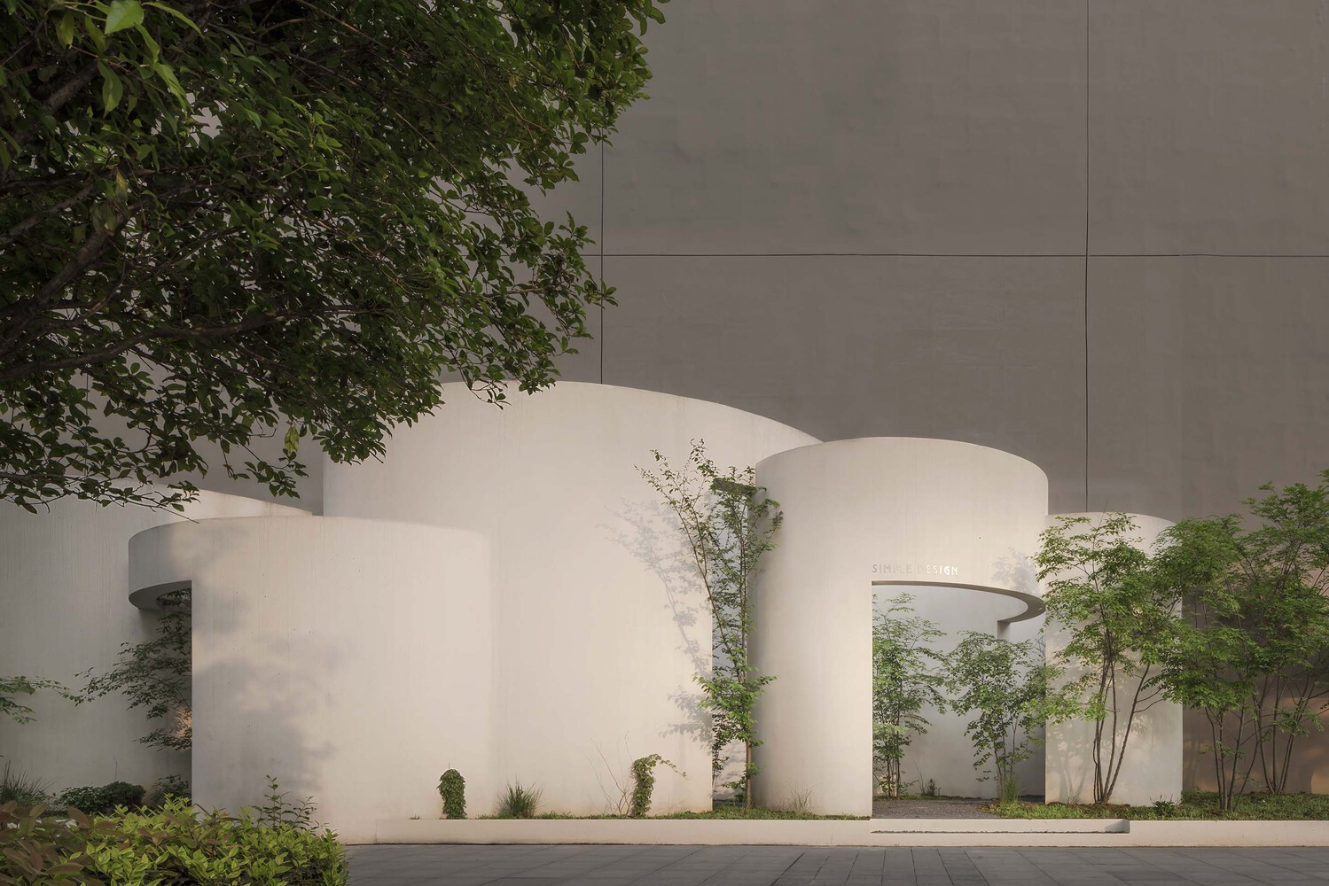White curved facade volumes with circular and arched openings interspersed with planted trees at twilight