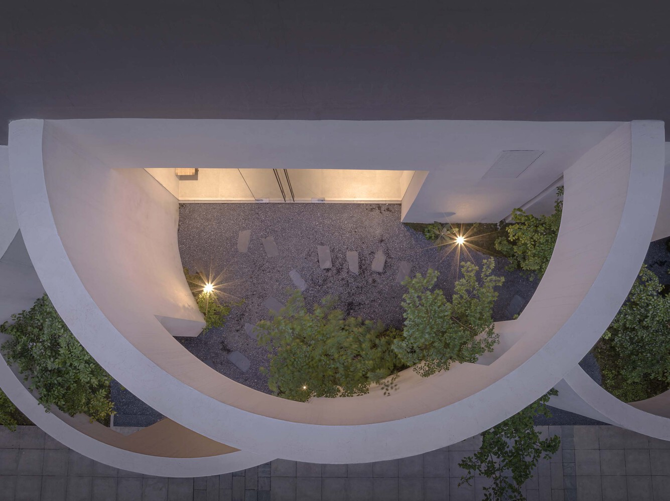 Aerial view of a curved courtyard with planted trees and gravel beds illuminated at twilight