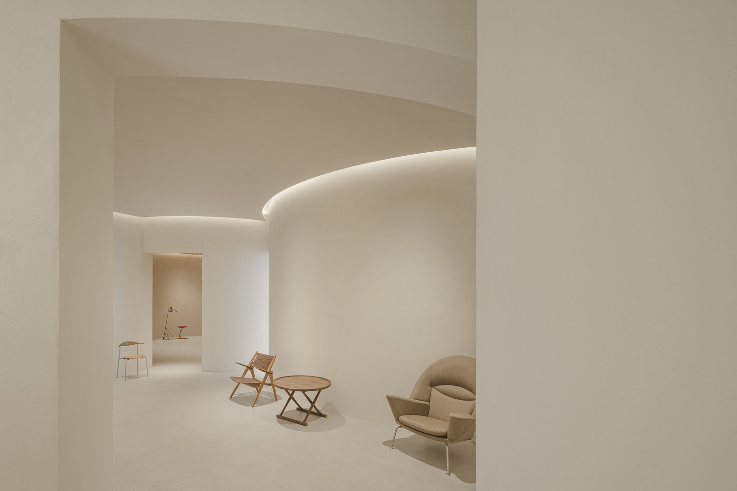 Gallery space with curved white walls, recessed cove lighting, and scattered timber furniture on pale flooring