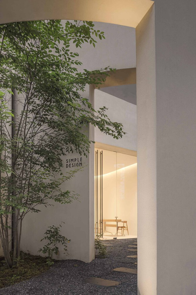 Courtyard entrance framed by white plaster walls with planted trees and gravel ground at dusk