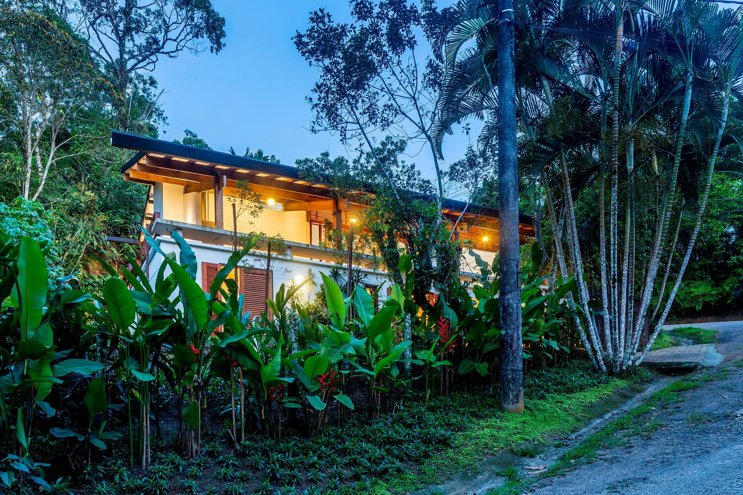 Two-story volume with overhanging timber roof nestled among tropical palms and banana plants at twilight