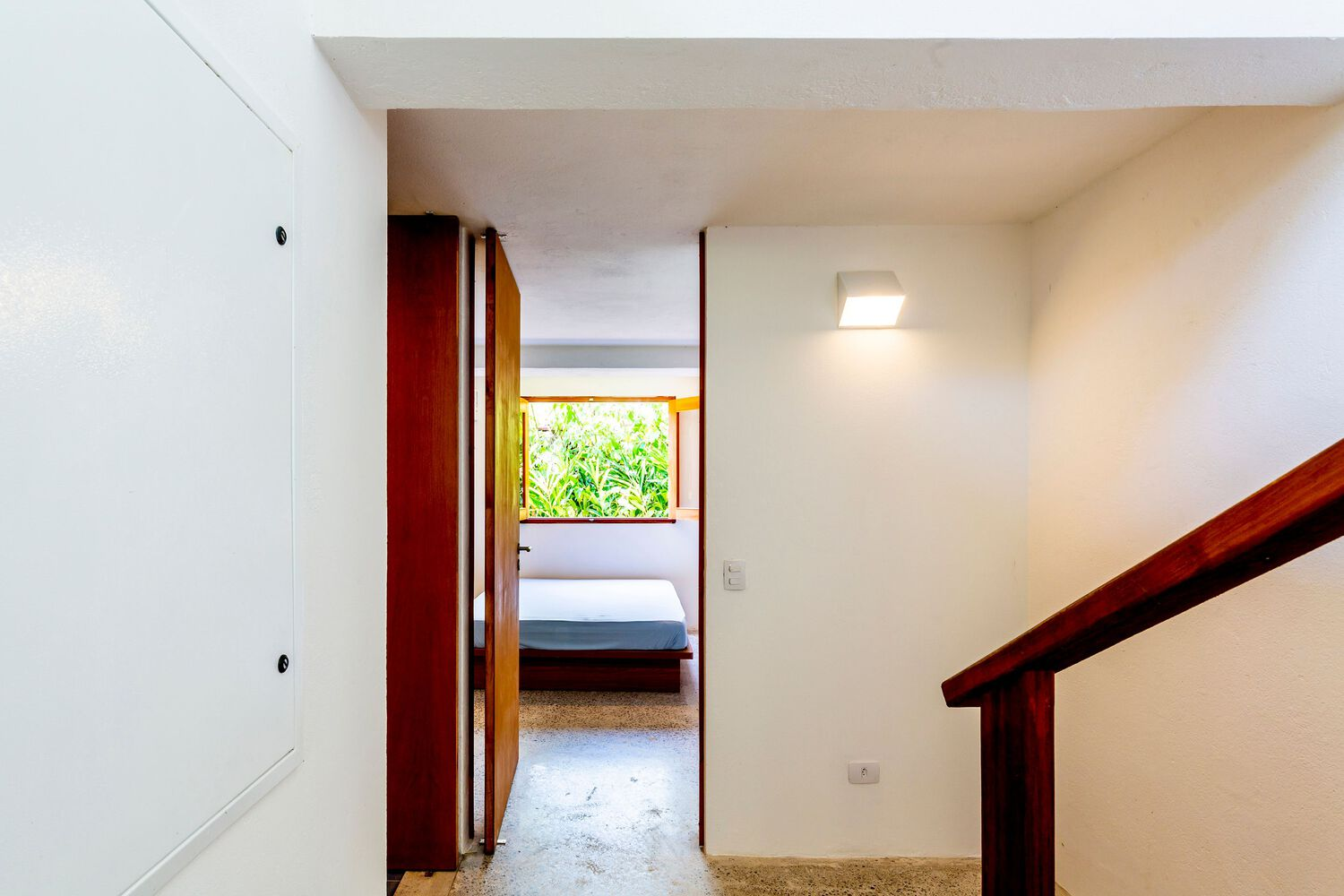 White interior hallway with timber handrail and deep red doorframe opening to a bedroom with terrazzo flooring