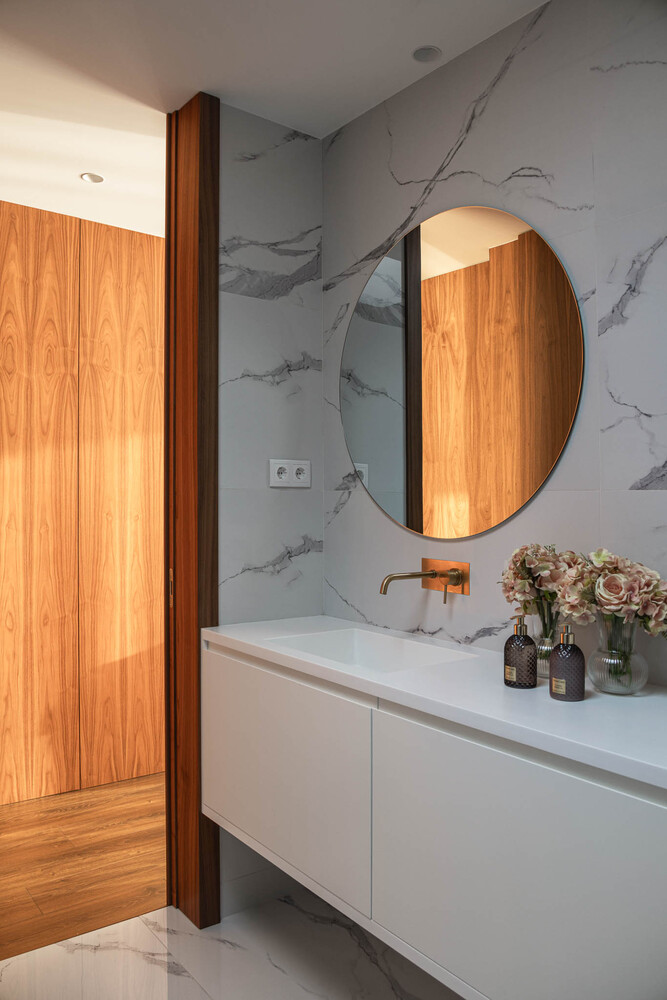 Bathroom vanity with white countertop and round mirror against grey veined marble wall with timber column