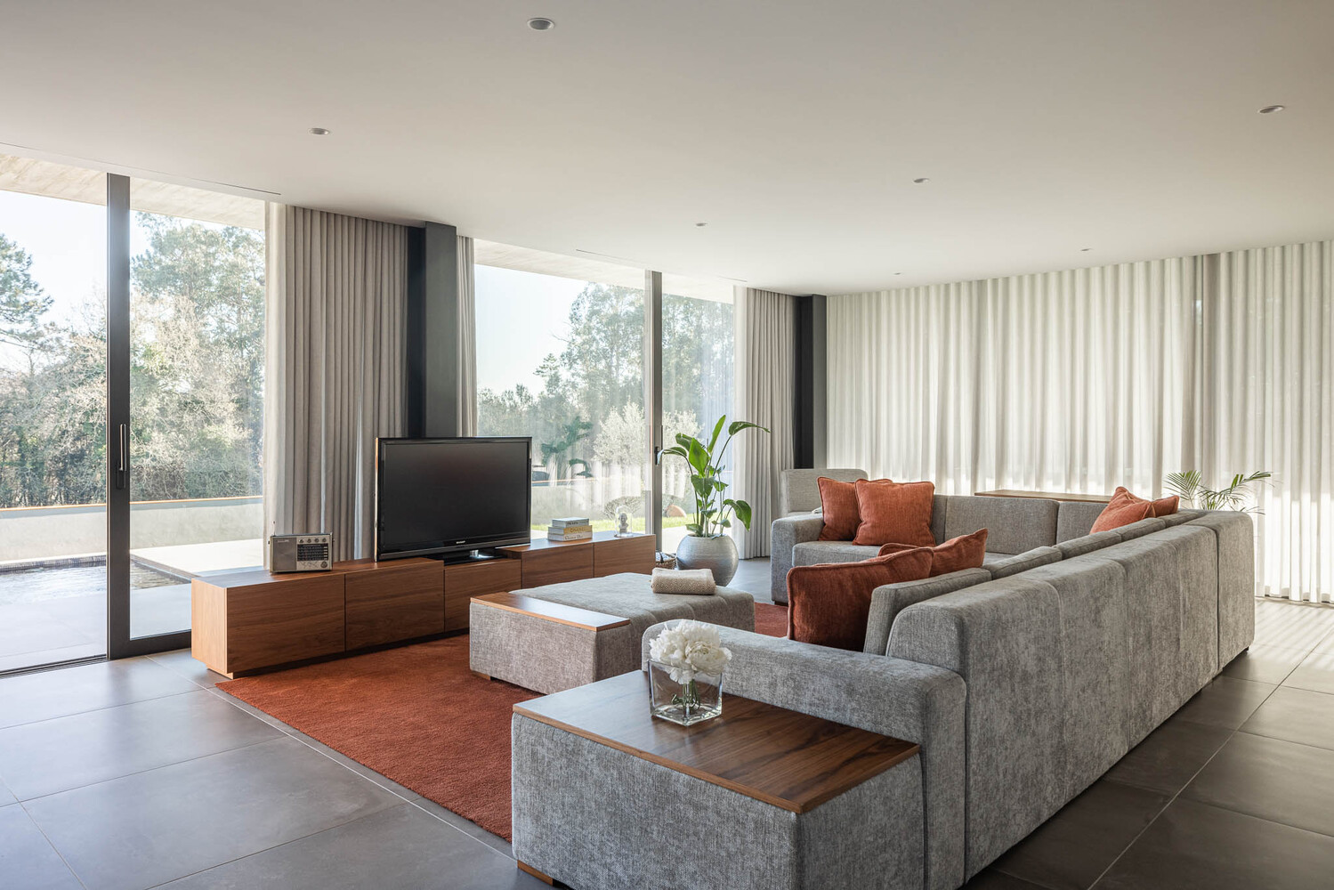 Living room with grey sectional sofa and floor-to-ceiling curtains framing glazed walls overlooking trees