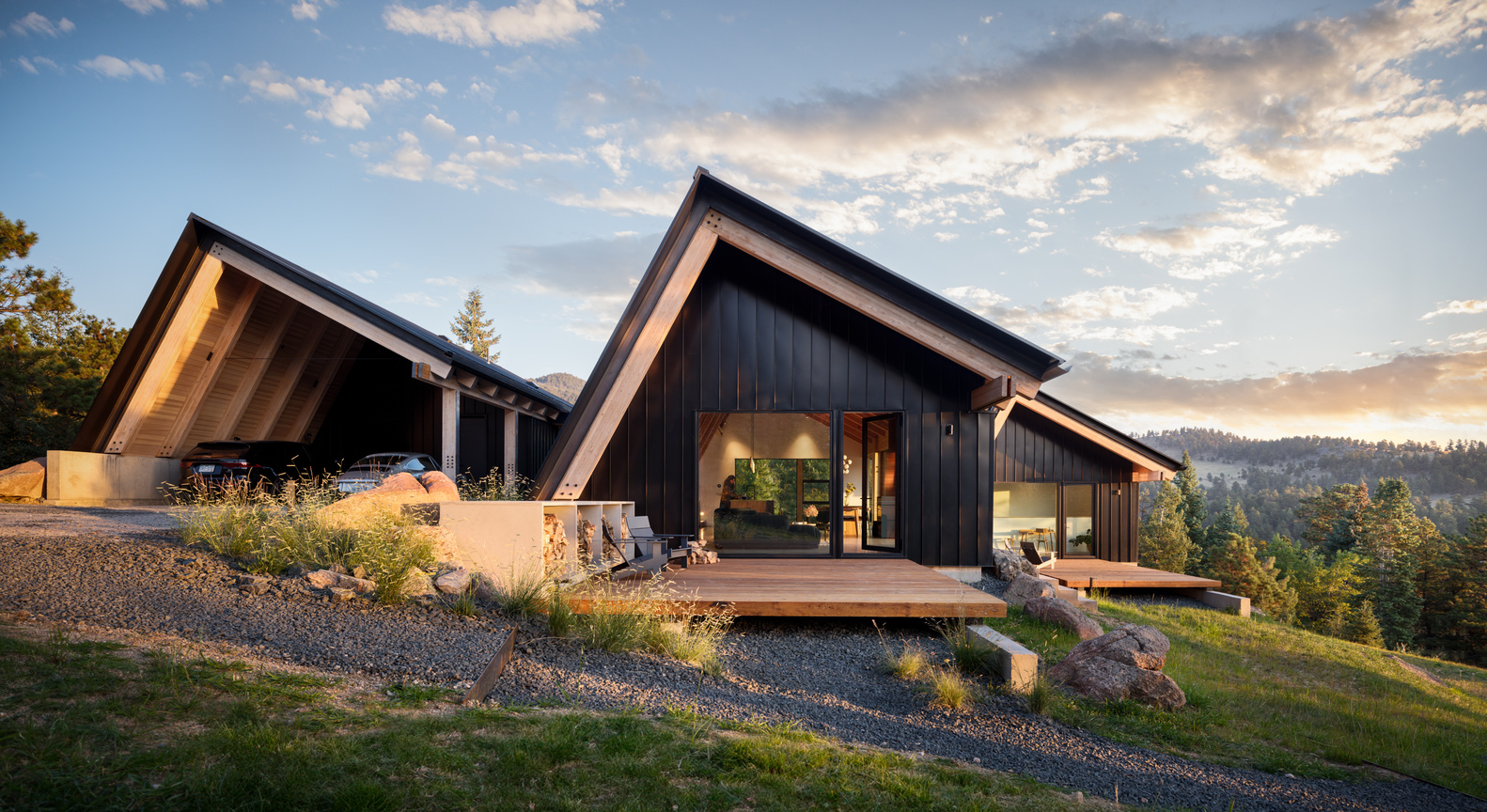 Two gabled volumes with dark metal cladding and timber-lined soffits on a gravel hillside at sunset
