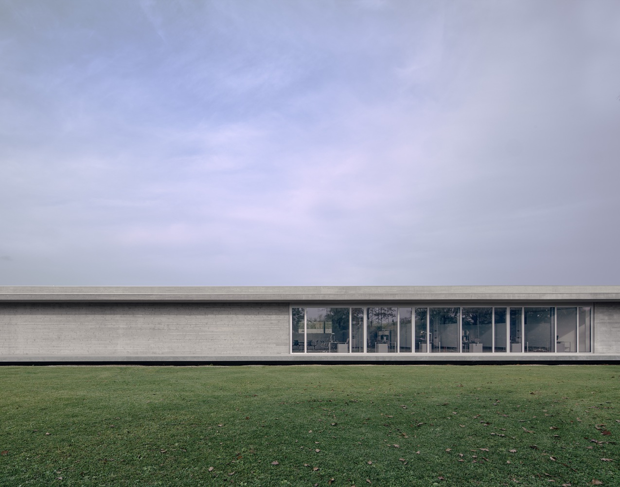 Long horizontal elevation with brick facade and continuous glazing across a lawn under overcast sky