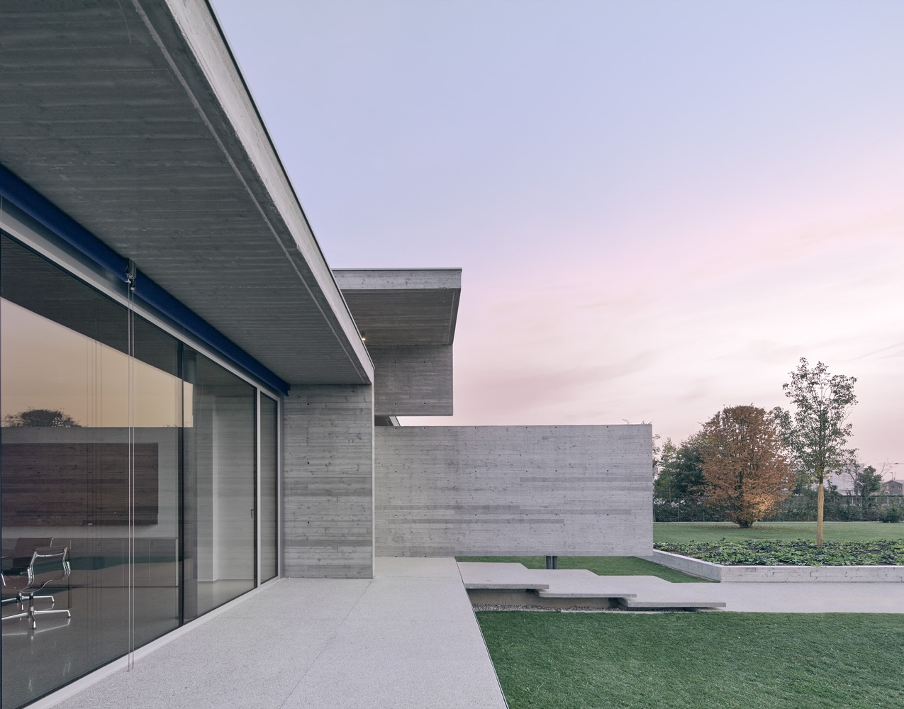 Covered terrace with board-formed concrete soffit and cantilevered volume overlooking lawn at dusk