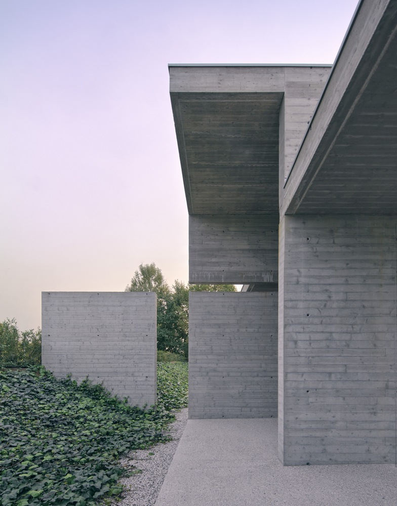 Covered walkway framed by board-formed concrete columns and cantilever with distant vegetation at dusk