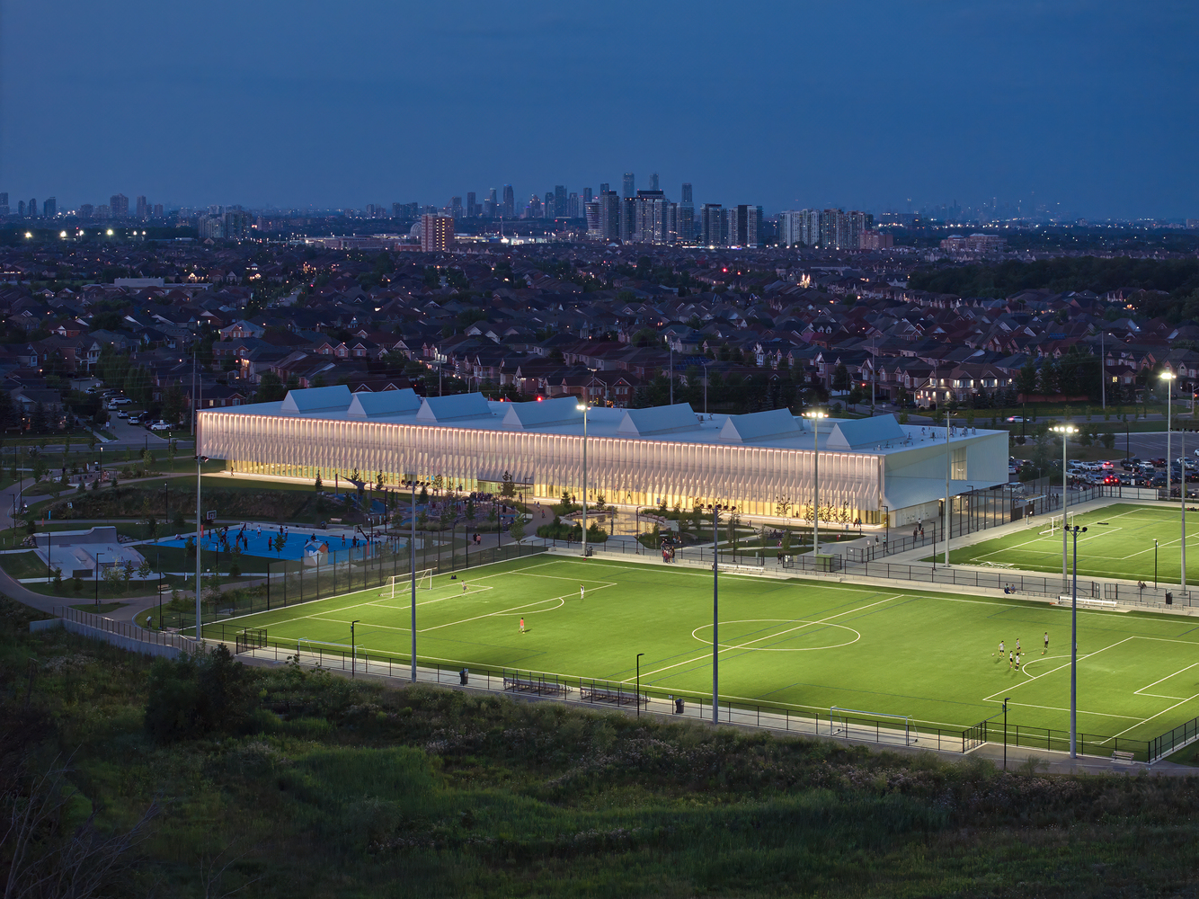 Illuminated sports facility at twilight overlooking soccer fields and distant residential skyline