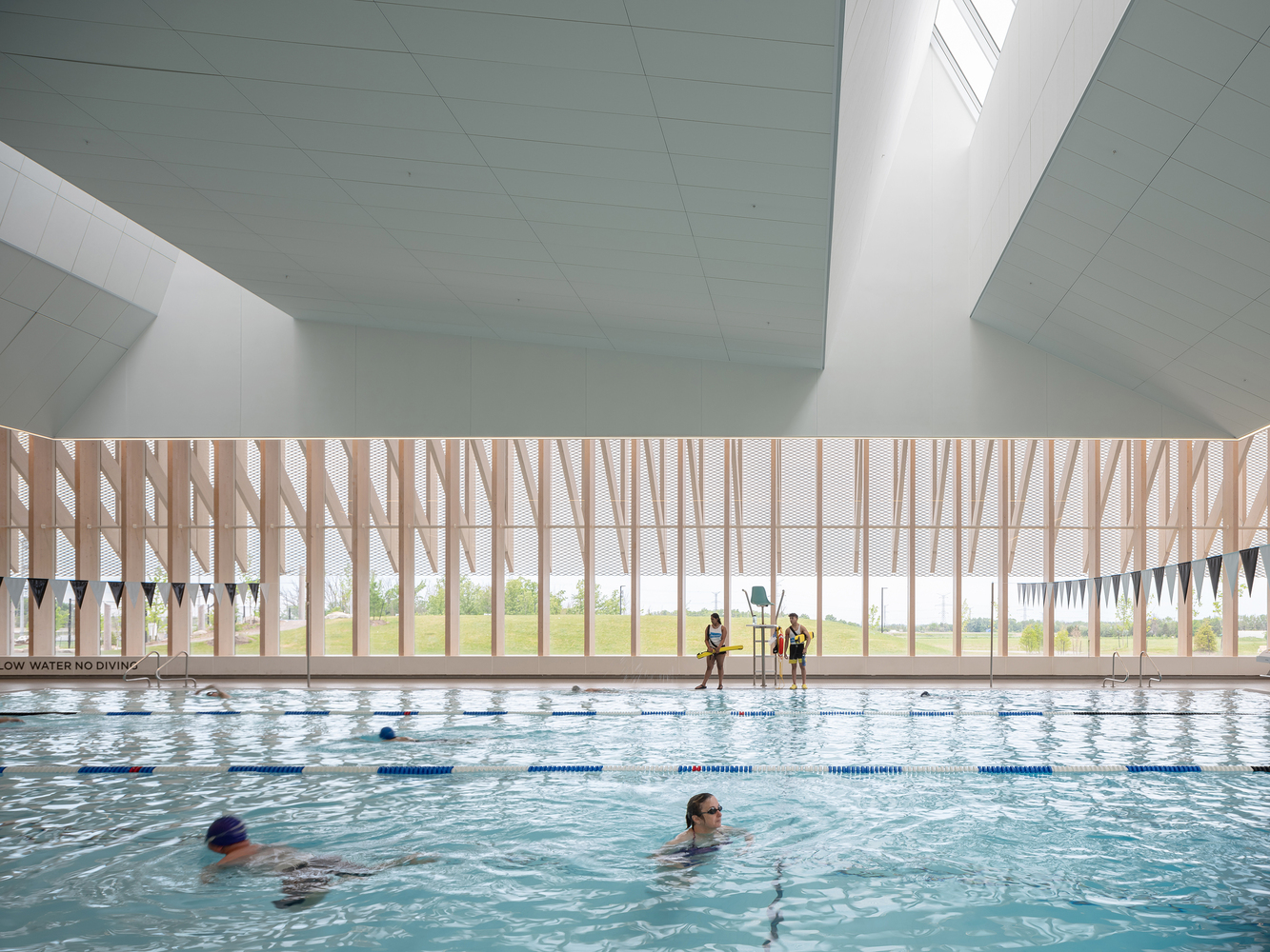 Indoor swimming pool with vertical timber louvre facade overlooking open fields and swimmers in the water