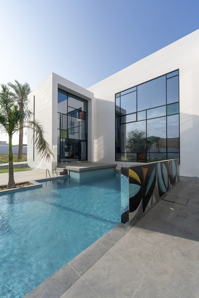 White stucco facade with floor-to-ceiling glass walls overlooking a blue pool with palm trees and grey paving