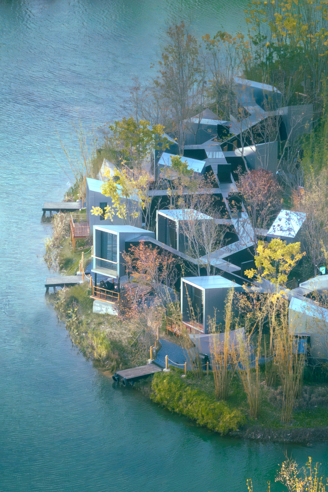 Drone view of scattered box-shaped volumes with white roofs connected by dark walkways among deciduous trees