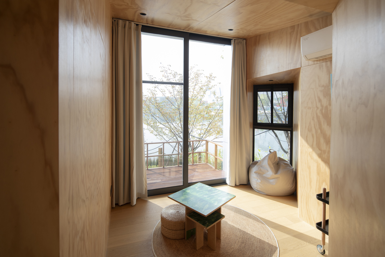 Plywood-lined room with window alcove overlooking lake and deck through floor-to-ceiling glass in afternoon light
